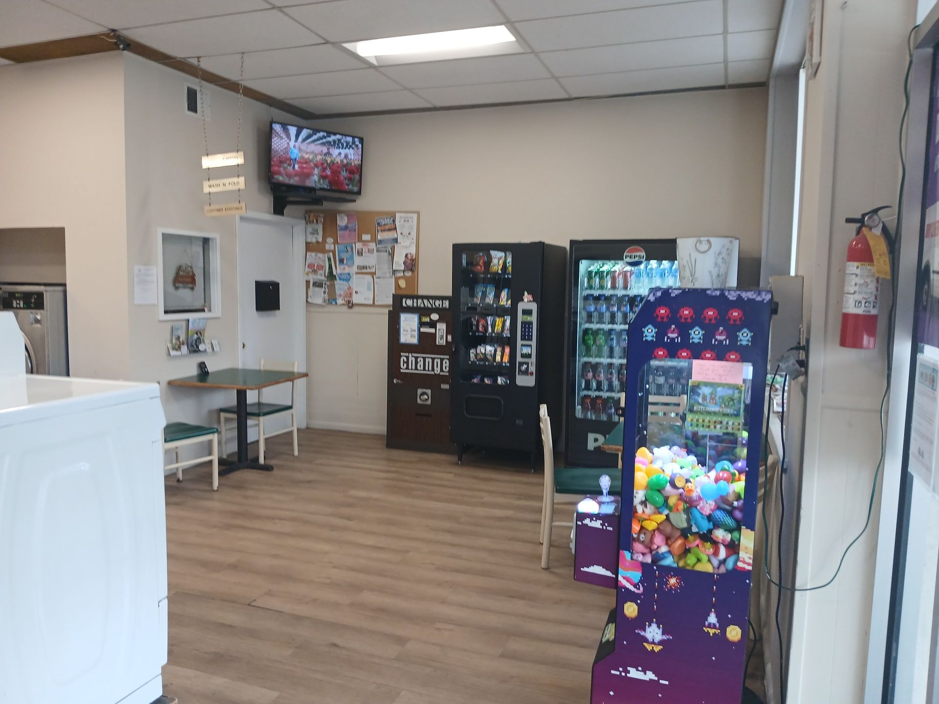 A small laundromat interior featuring a vending machine, a claw prize machine, a television, and a small table with chairs.