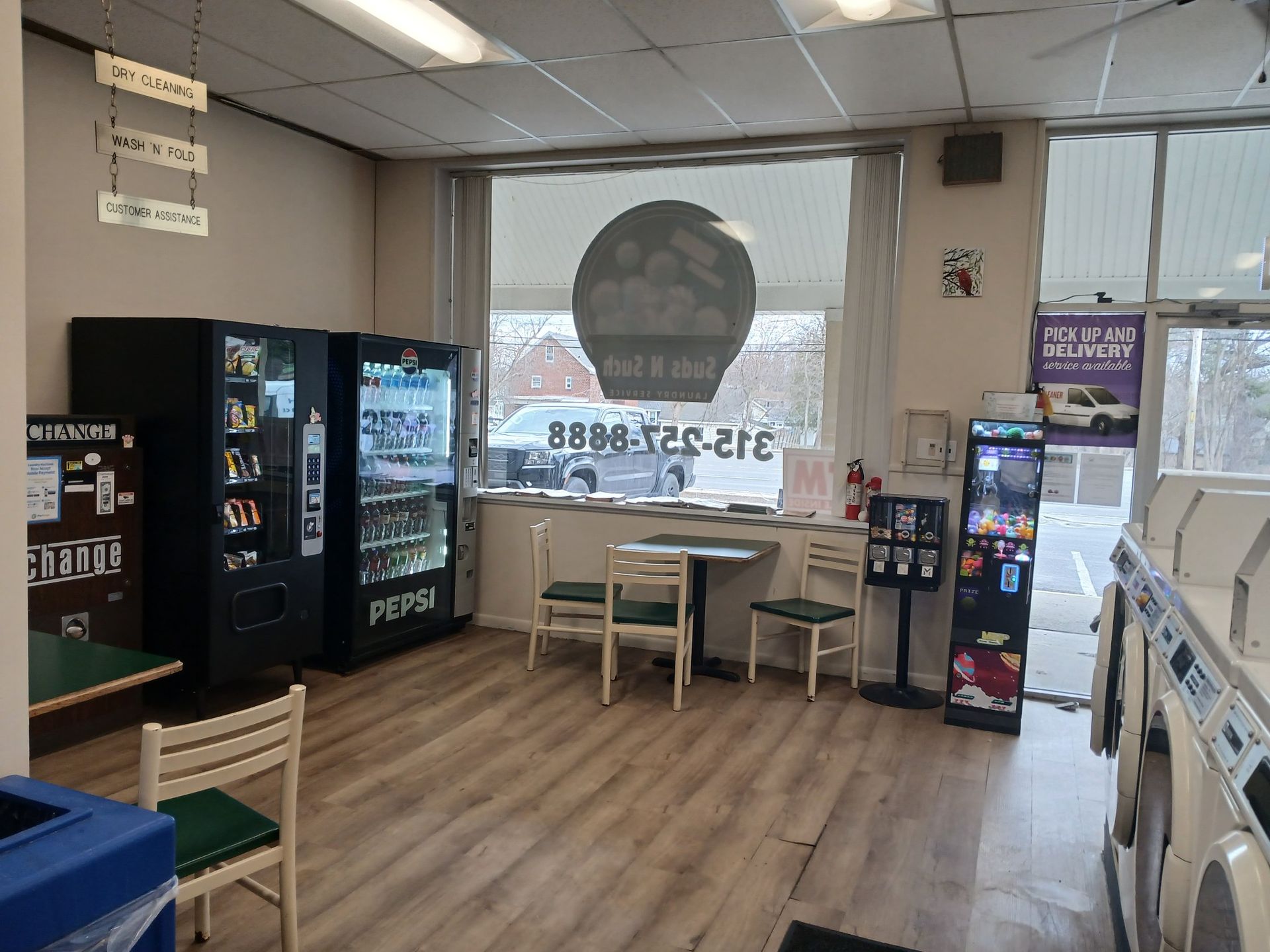 A laundromat interior featuring vending machines, a small dining area, and a row of washing machines along the wall.