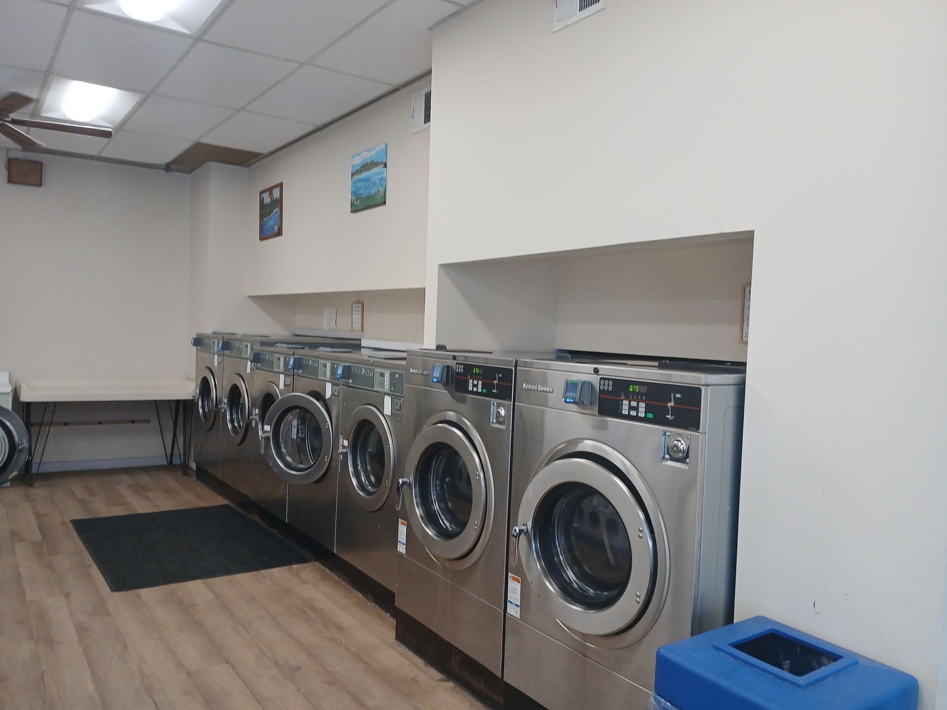 A row of stainless steel dryers inside a laundromat with wood-look flooring and white walls.