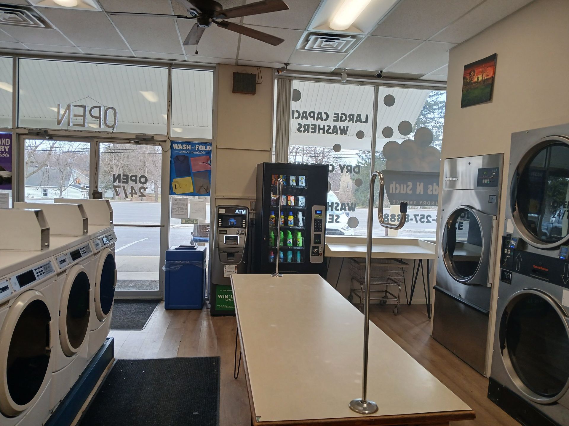 A laundromat interior with a folding table in the center, rows of front-load washers, and a vending machine.