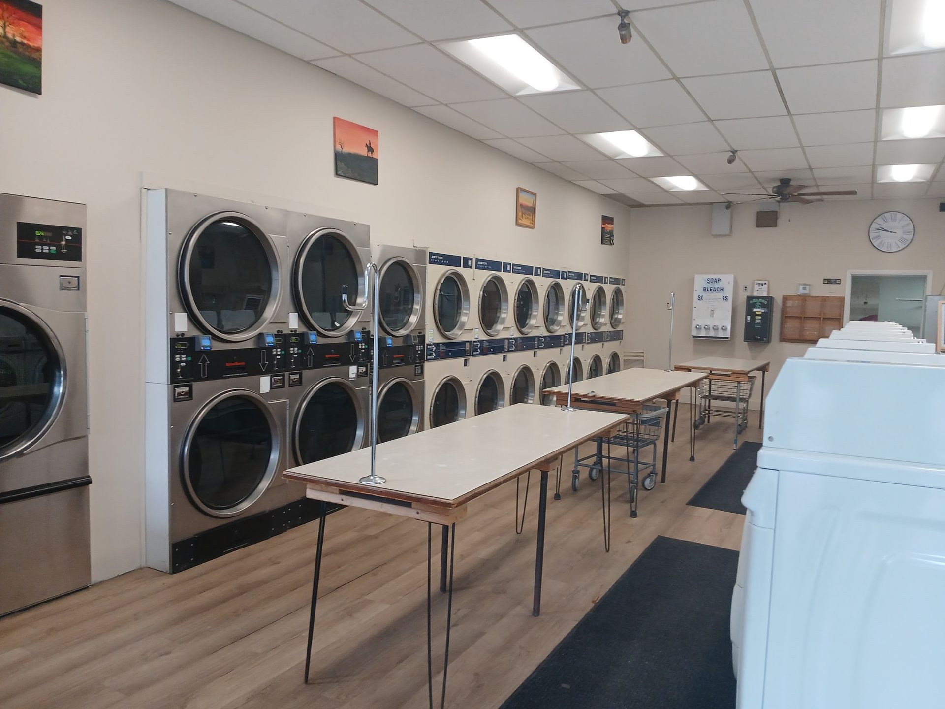 Empty interior of a laundromat with rows of stainless steel dryers, folding tables, and light-colored flooring.