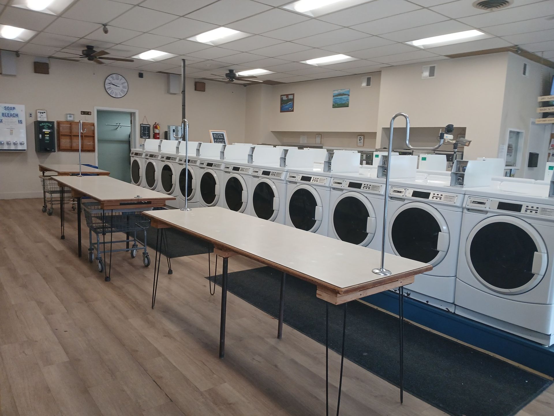 A row of white clothes dryers in a laundromat with two folding tables in the foreground.