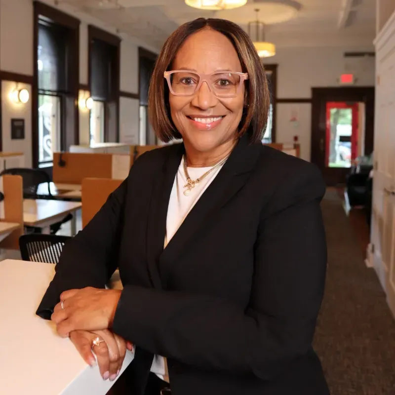 Woman in blazer and glasses smiles, leaning on a white counter in a modern office space.