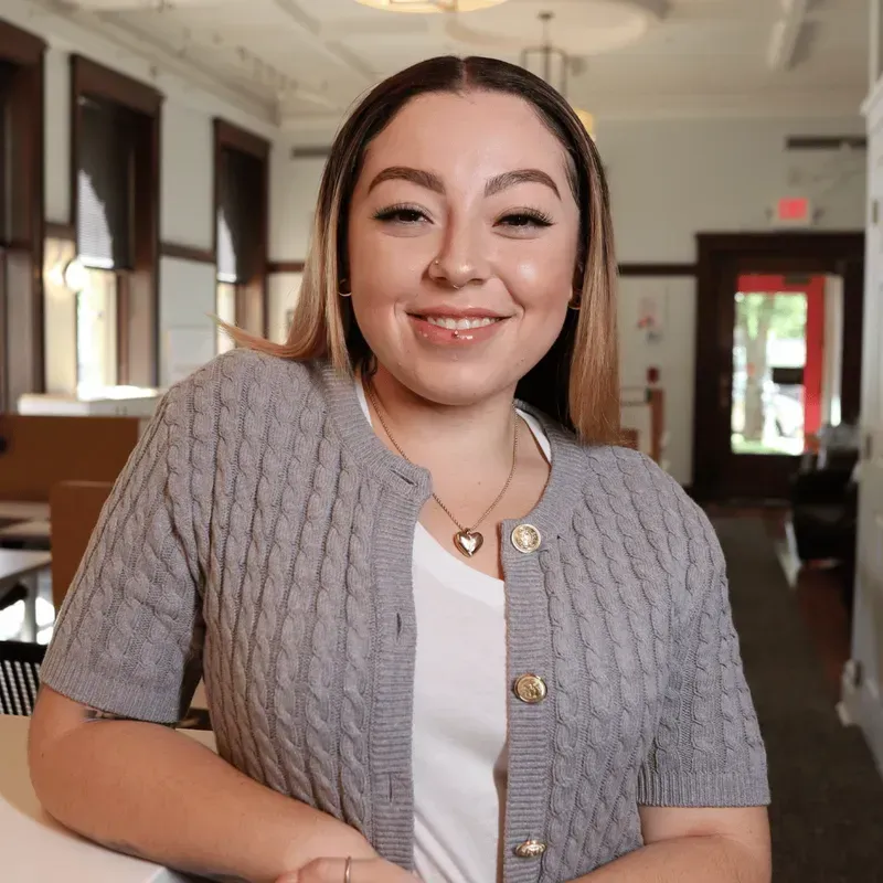 Woman in gray cardigan smiles indoors; a library or office setting.