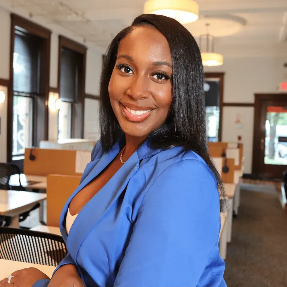 Woman in blue blazer smiles, seated in a well-lit office.
