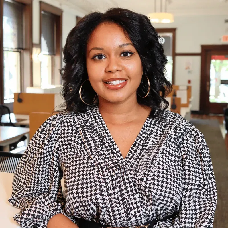 Woman in a black and white houndstooth top, smiling, in an office setting with natural light.