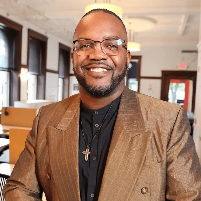 Man wearing glasses and a blazer smiles in a bright office space.