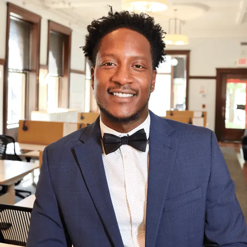 Man in a blue suit with a black bow tie smiles in an office setting.