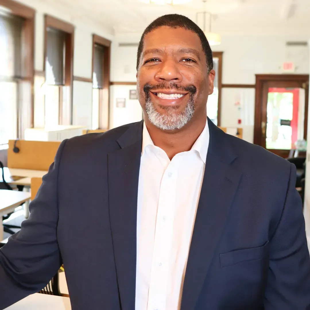 Man in a navy suit smiles at the camera in an office setting.