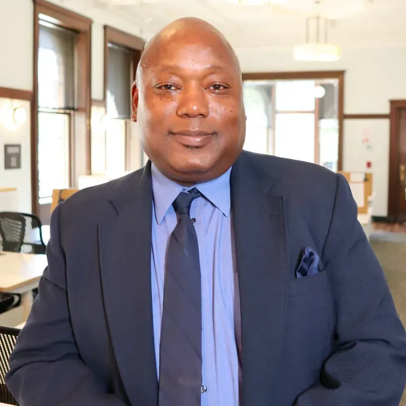 Man in a suit smiles, inside a building with windows and open space.