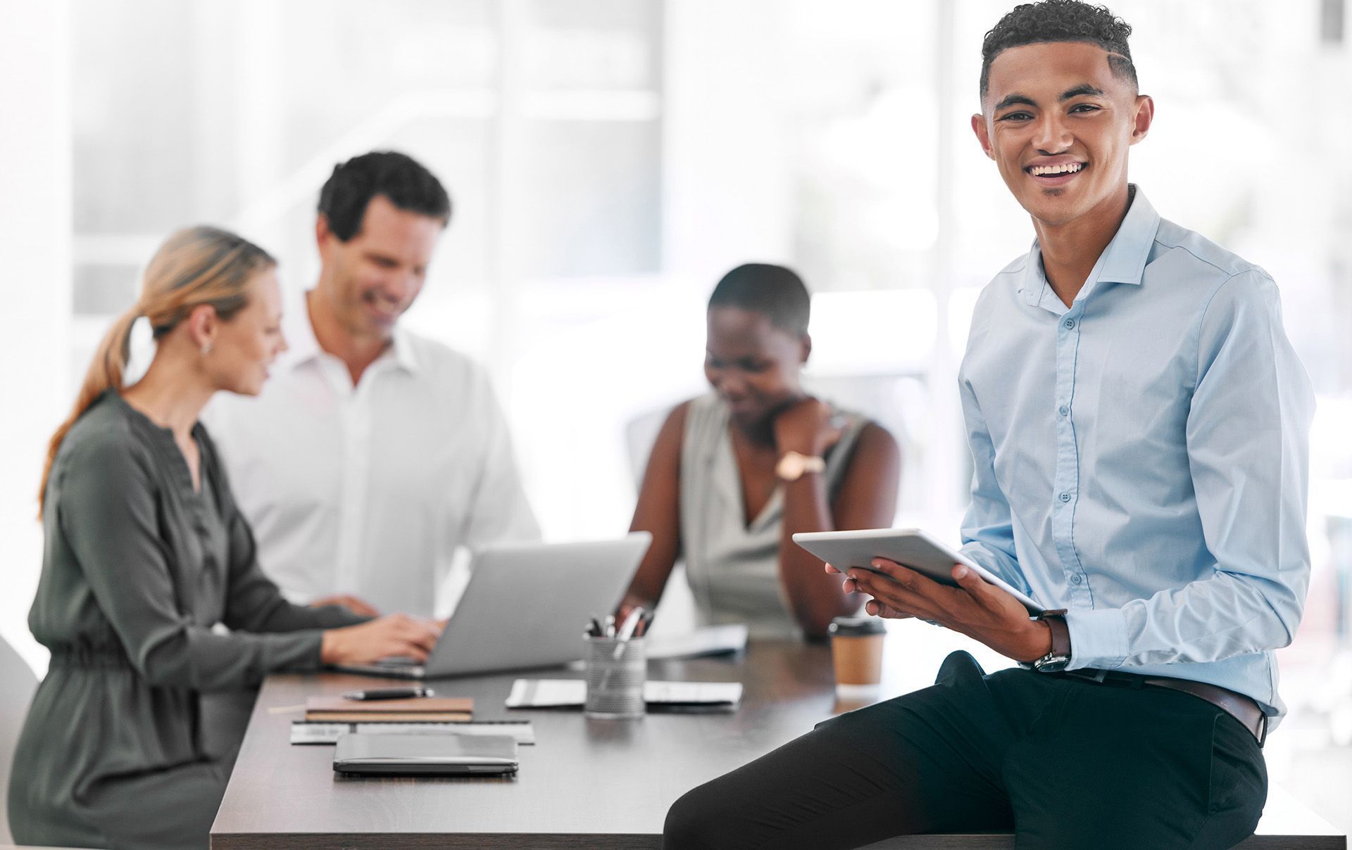 Man holding tablet smiles, leans against a table with colleagues working on laptops in an office.