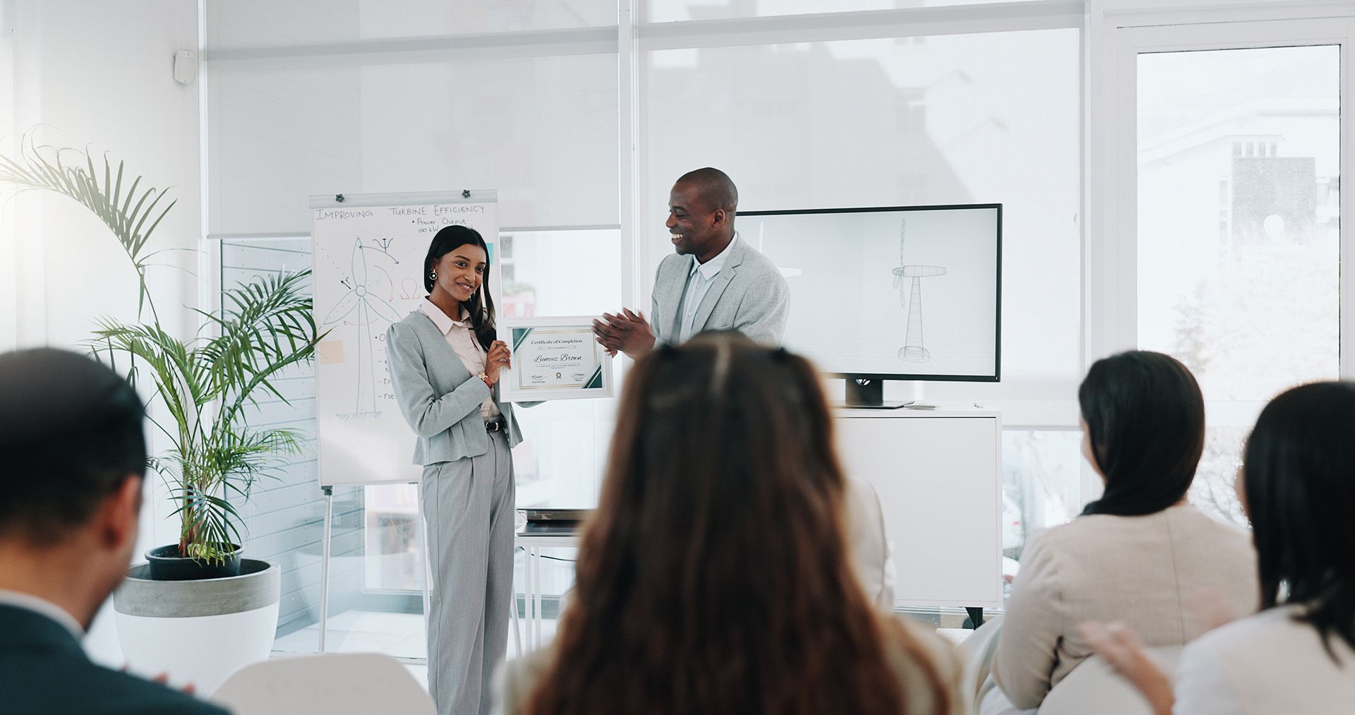 Two people presenting in front of an audience, a whiteboard, and a screen in a bright office.