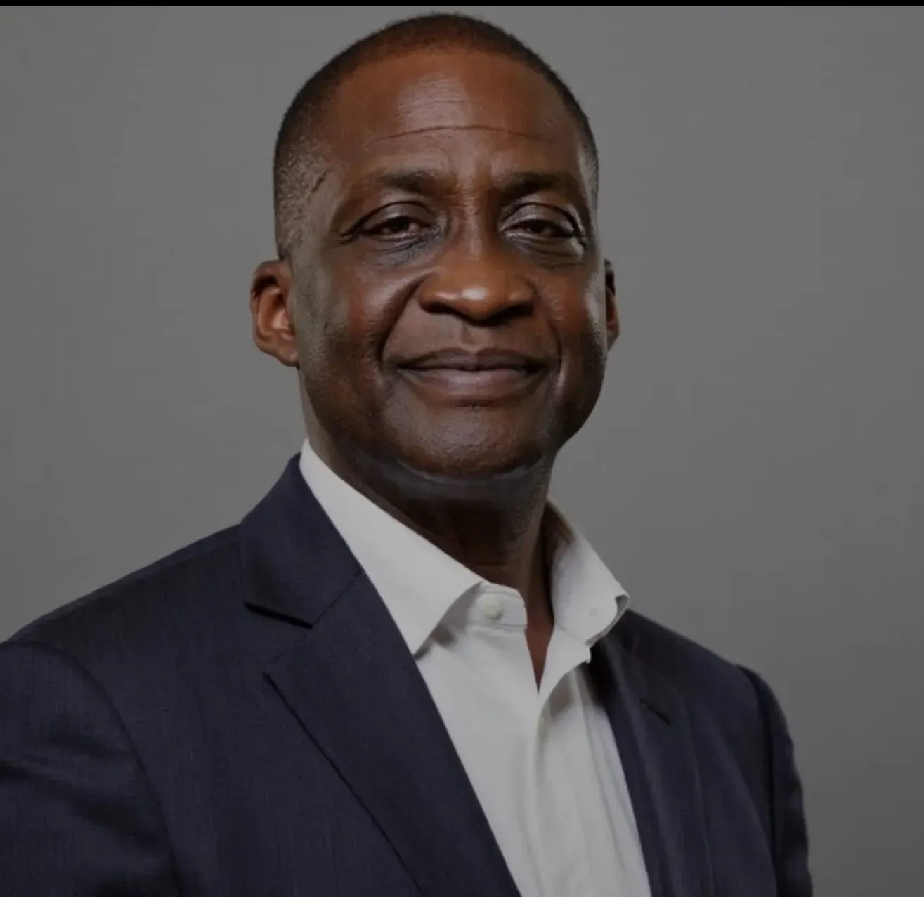 Man in navy suit and white shirt, smiling, against a gray background.