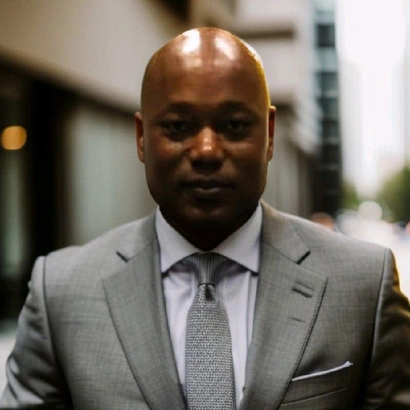 Man in a suit smiles, inside a building with windows and open space.