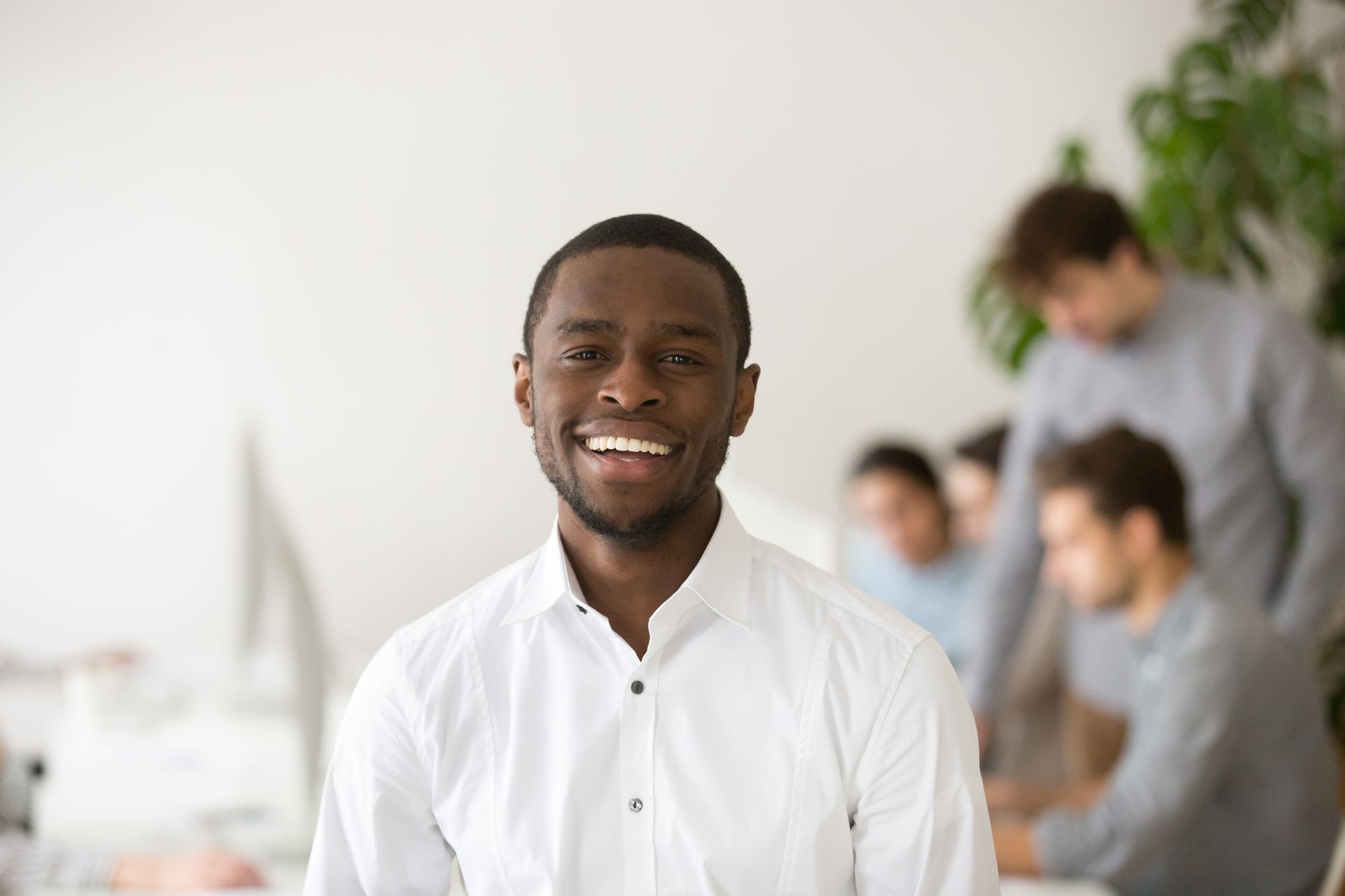 Man in white shirt smiles directly at the viewer with colleagues in the blurred background of an office setting.