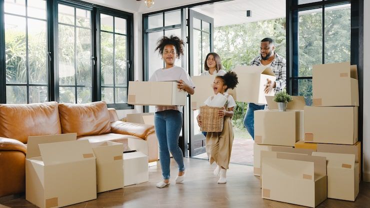Family carrying moving boxes into a new home with windows and a sofa.