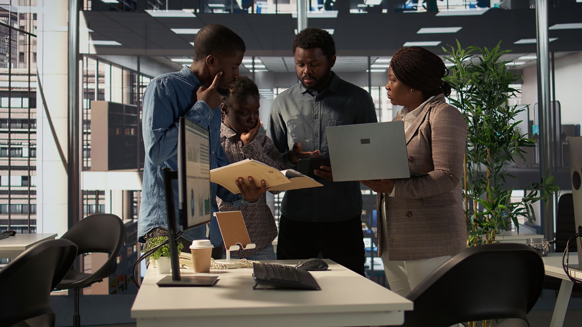 Four people in office looking at documents, one with a laptop, by a window.