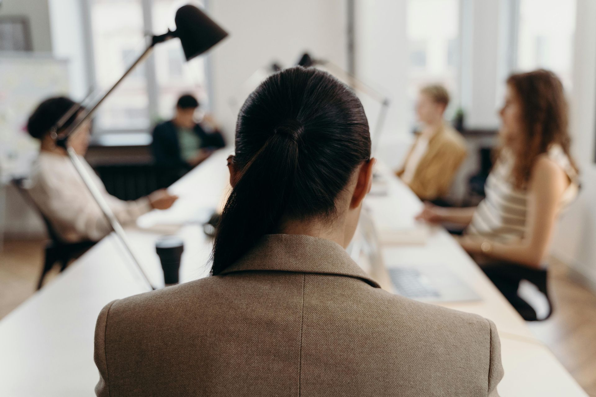 Woman in tan blazer at a conference table, facing blurred colleagues, lamp overhead.