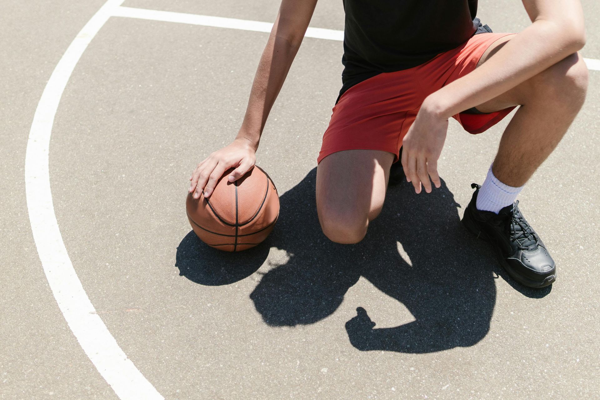 Person kneeling on basketball court, hand on ball. Red shorts, black shoes, shadow.