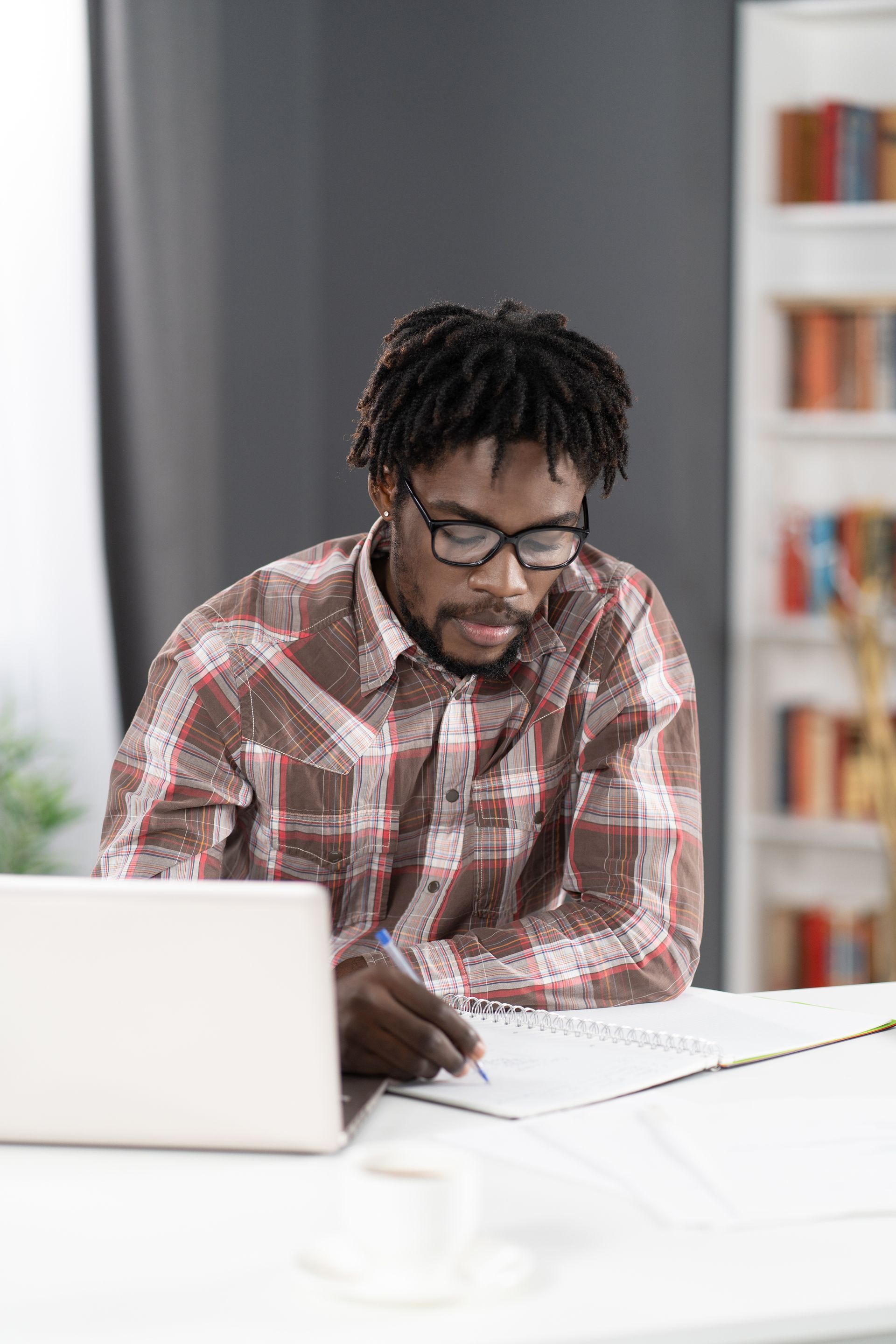 Man writing in a notebook, looking at a laptop. Wearing glasses and plaid shirt, at a desk with coffee.