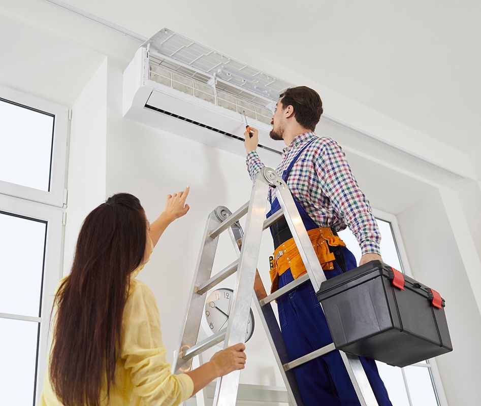 A man is standing on a ladder fixing an air conditioner while a woman looks on.