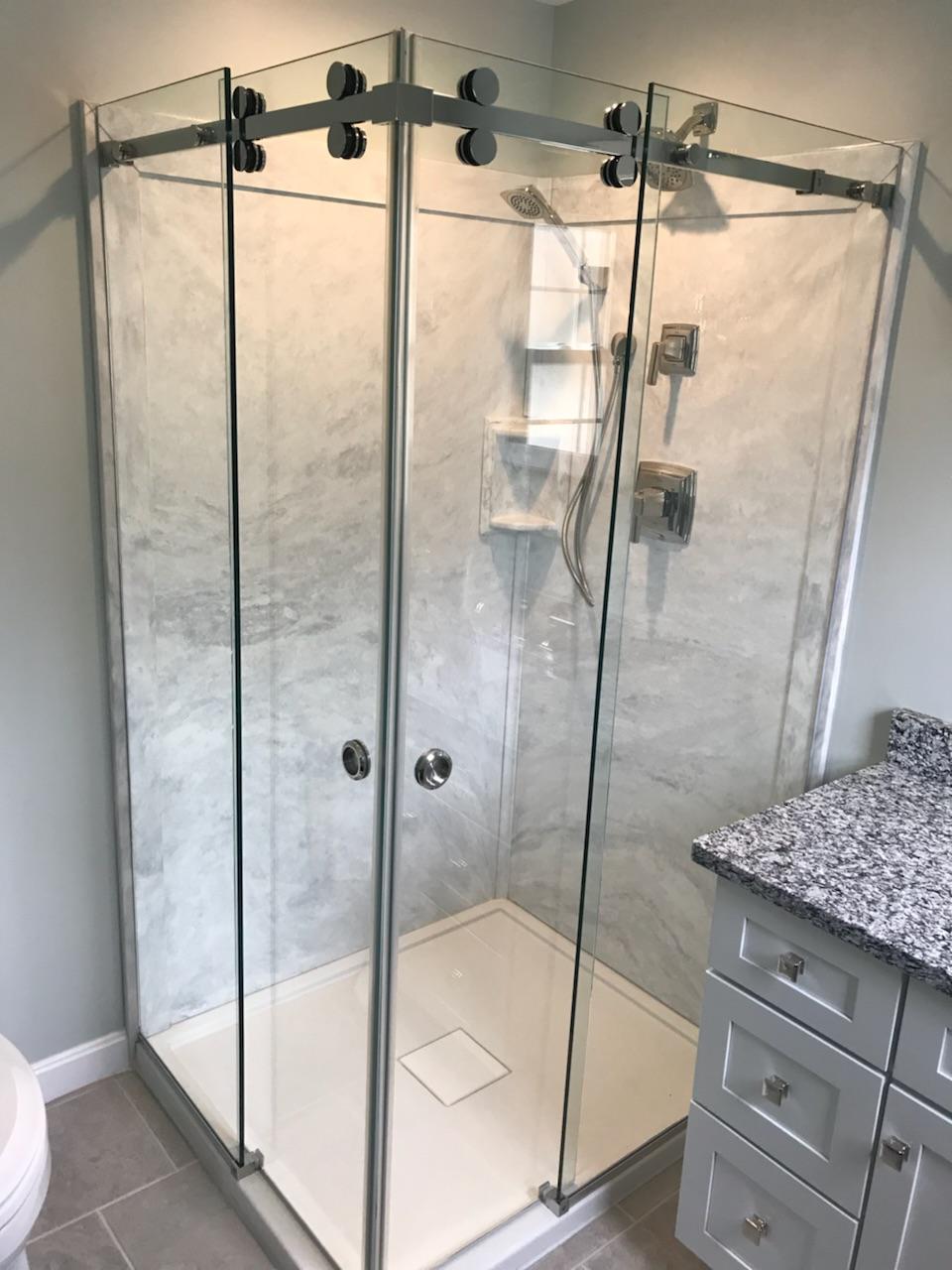 Corner shower with clear glass doors, marble-look walls, and a white base, beside a gray vanity with granite countertop.