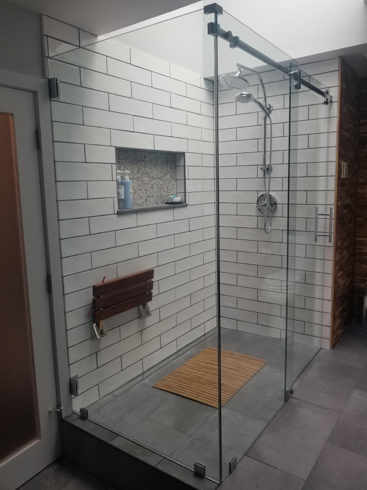 Modern bathroom with a glass shower, white brick tile, wooden seat, and gray floor.