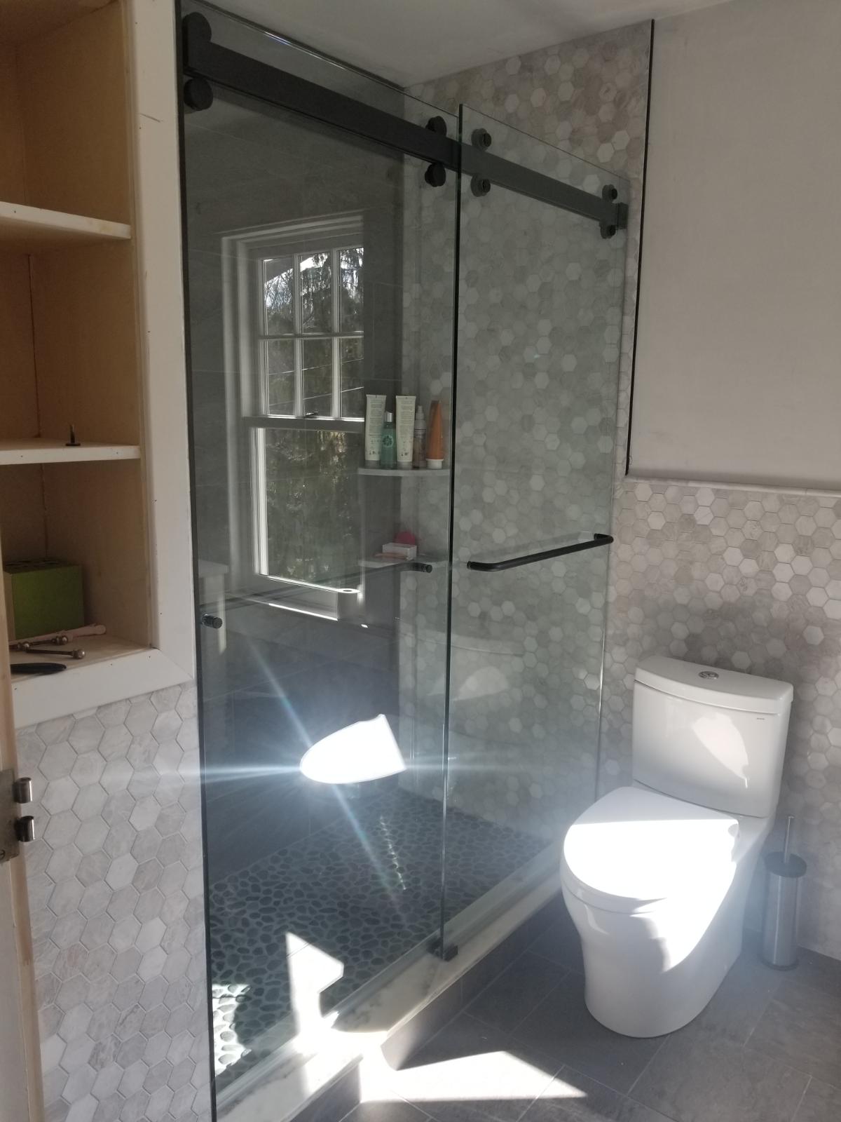 Bathroom with sliding glass shower, toilet, and built-in cabinet. Gray hexagon tile and black hardware.