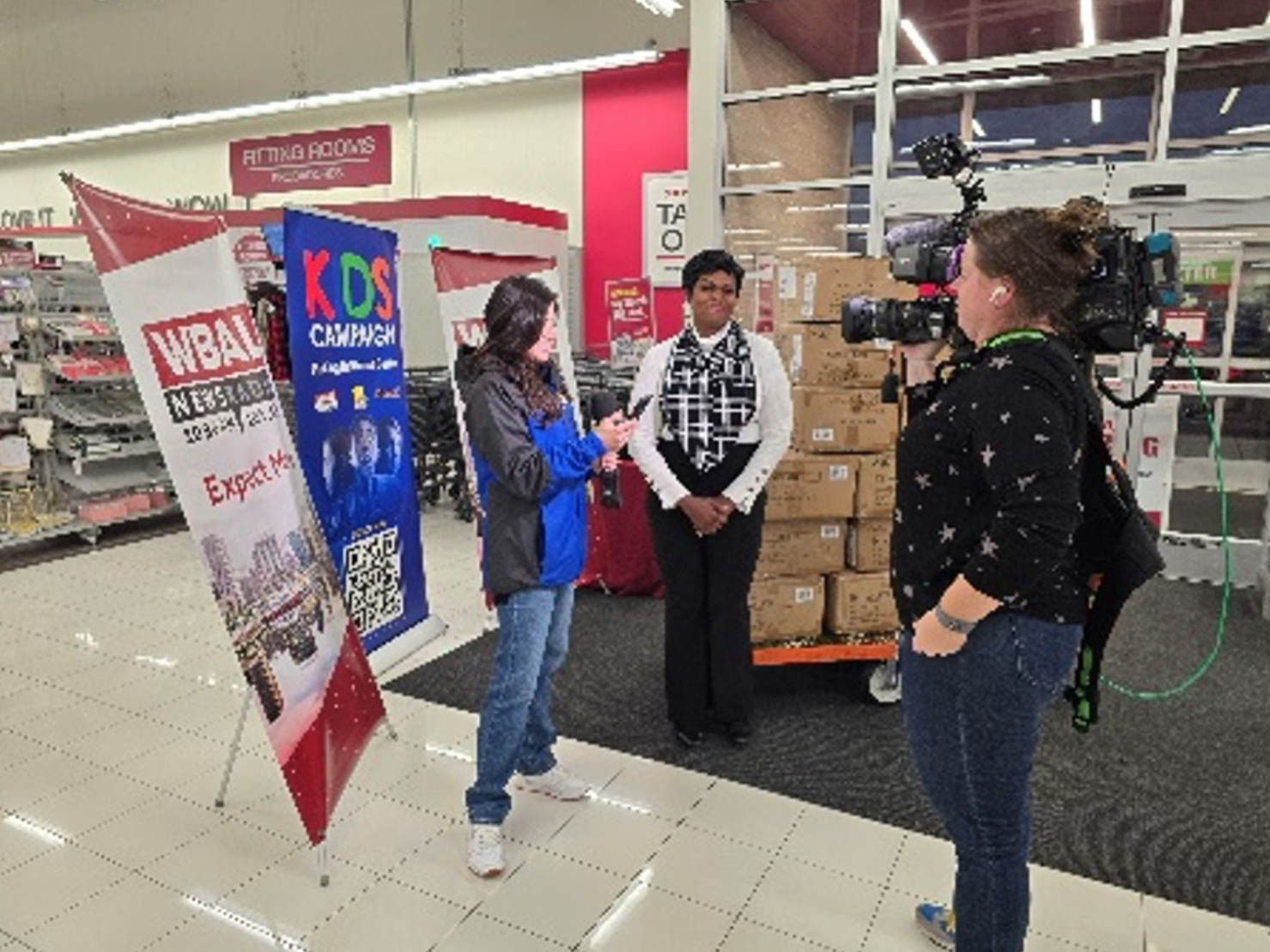 News crew interviews a woman in a store; boxes on a cart in the background, banner on the left.
