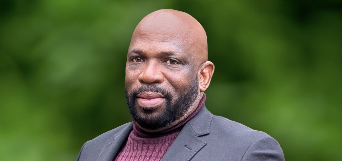 A man in a suit and sweater is standing in front of a green background.