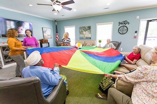 A group of elderly people are playing with a colorful parachute in a living room.