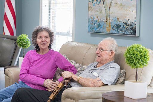 An elderly couple is sitting on a couch in a living room holding hands.