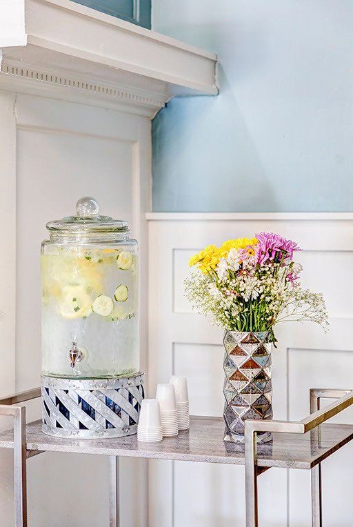 A drink dispenser and a vase of flowers are on a table.