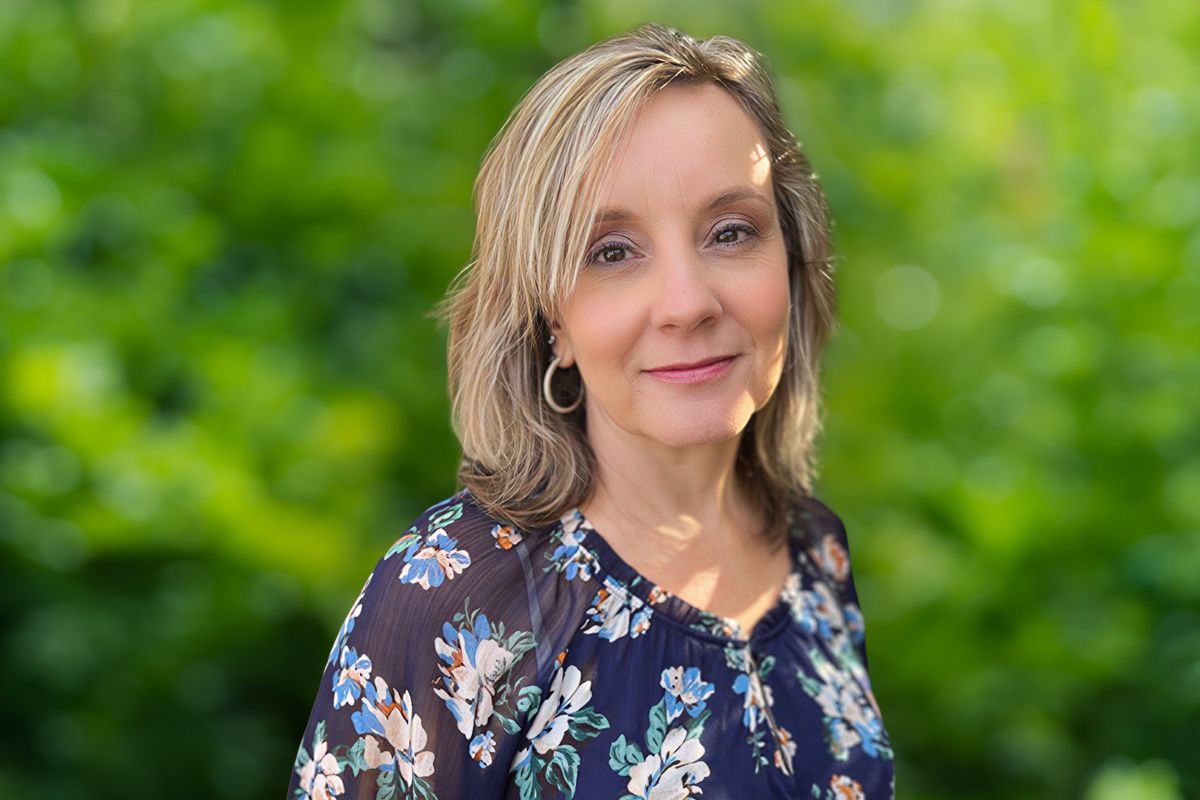 Woman with blonde hair, wearing a floral blouse, smiles against a bright background.