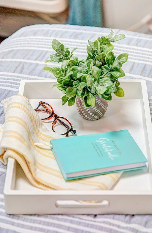 A white tray with a book , glasses , and a plant on a bed.