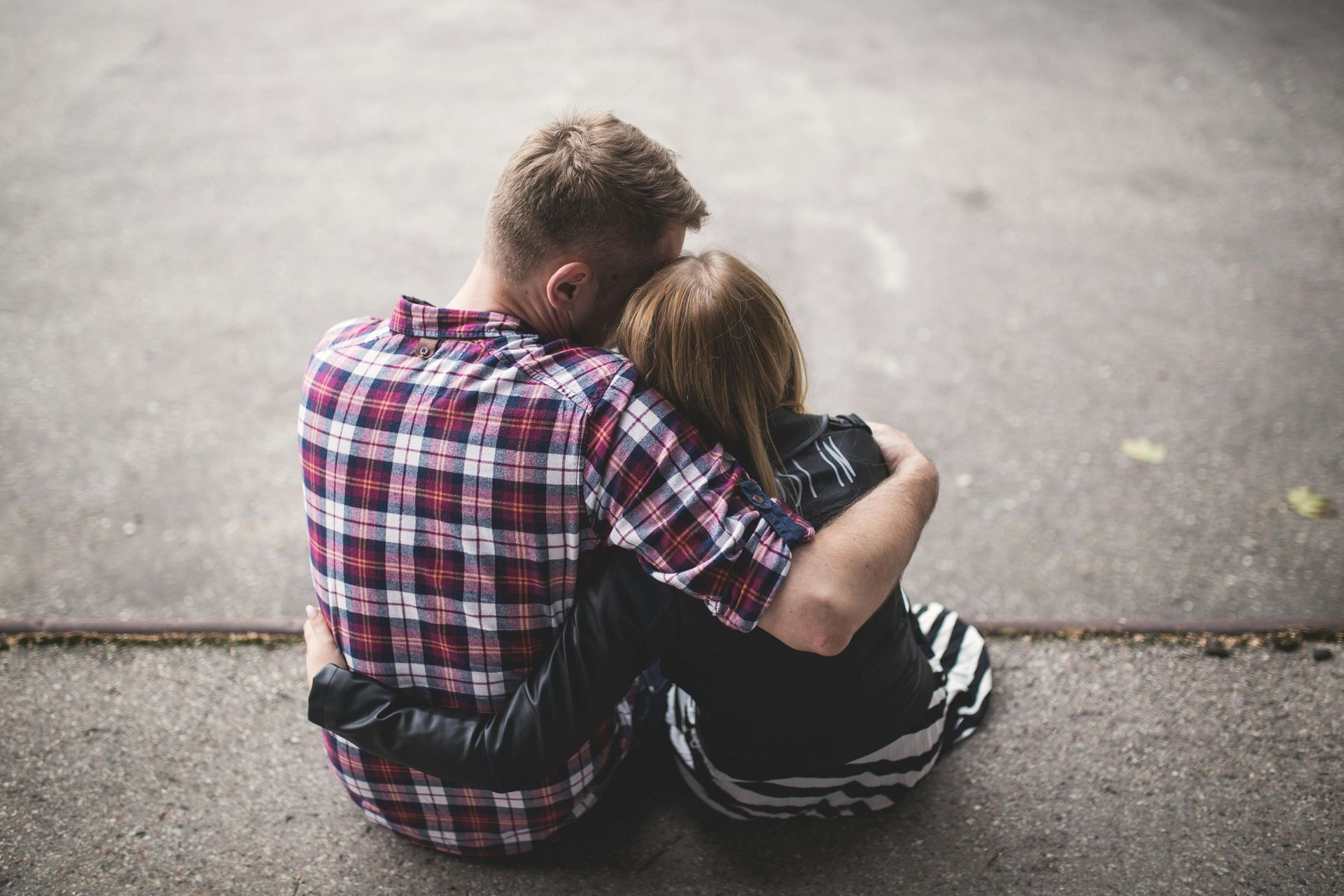 Man in plaid shirt hugging a person with brown hair and a striped skirt on concrete.