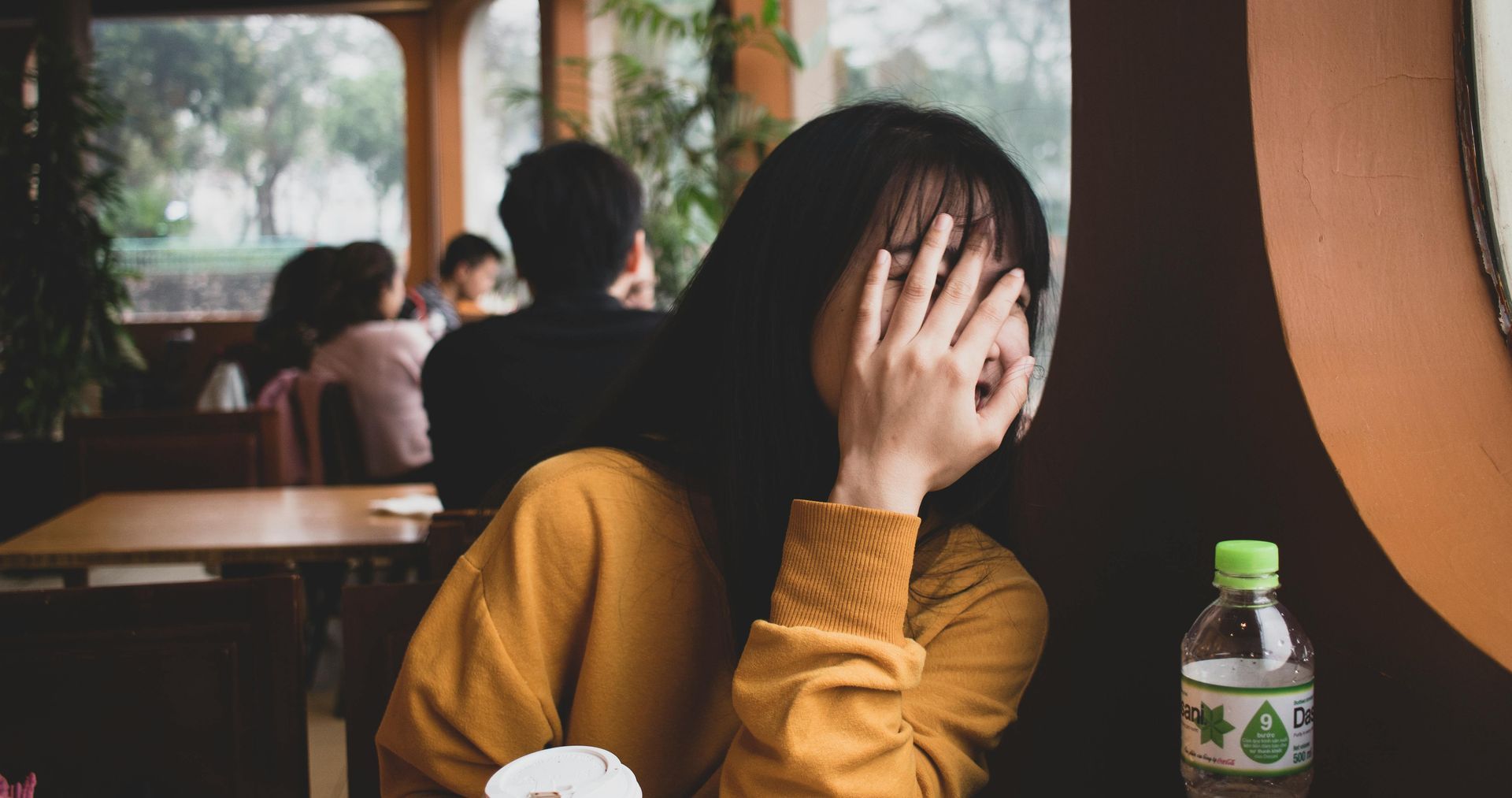 Woman in yellow sweater covers face at cafe table, obscured background.