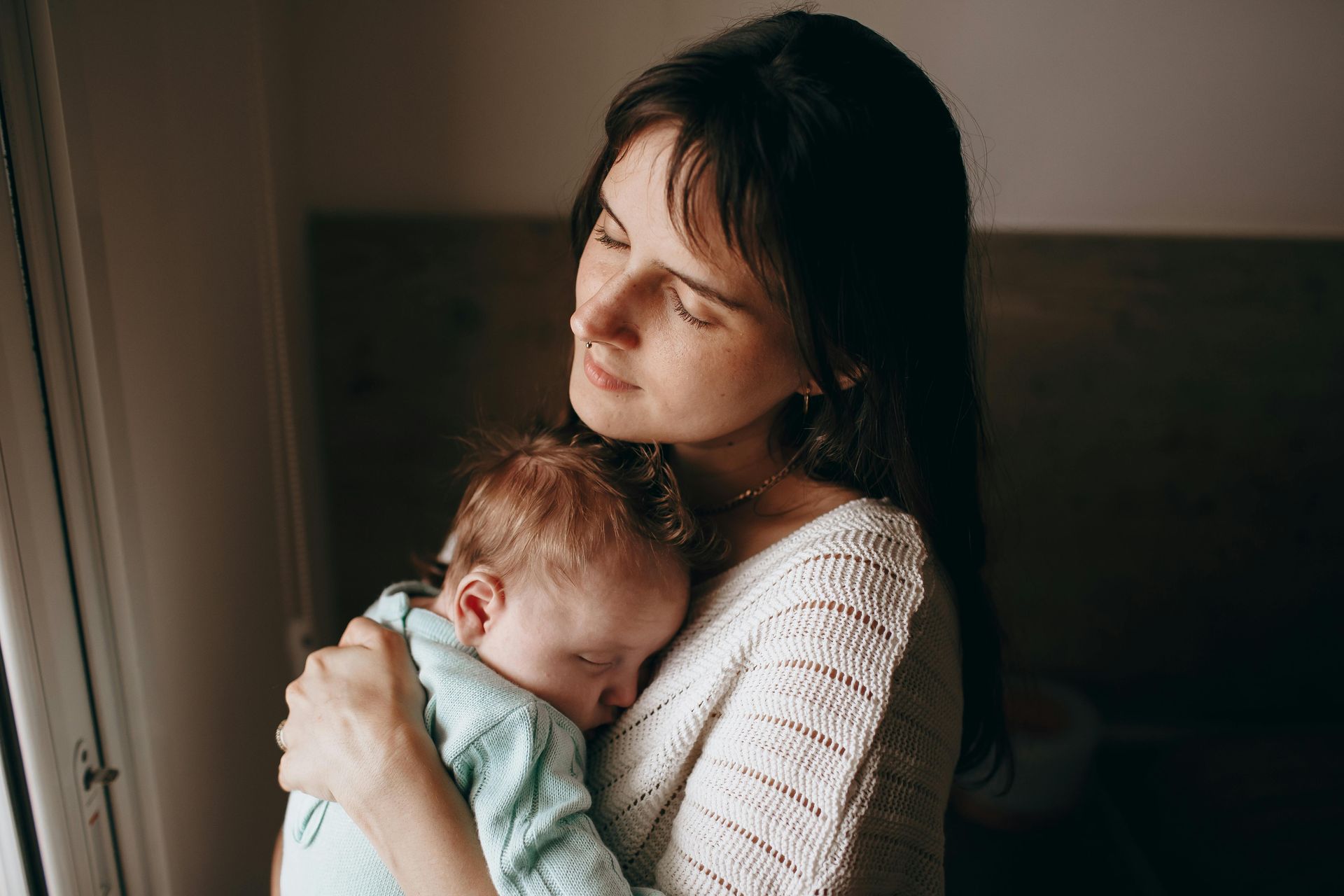 Woman holding a sleeping baby; both bathed in sunlight near a window.