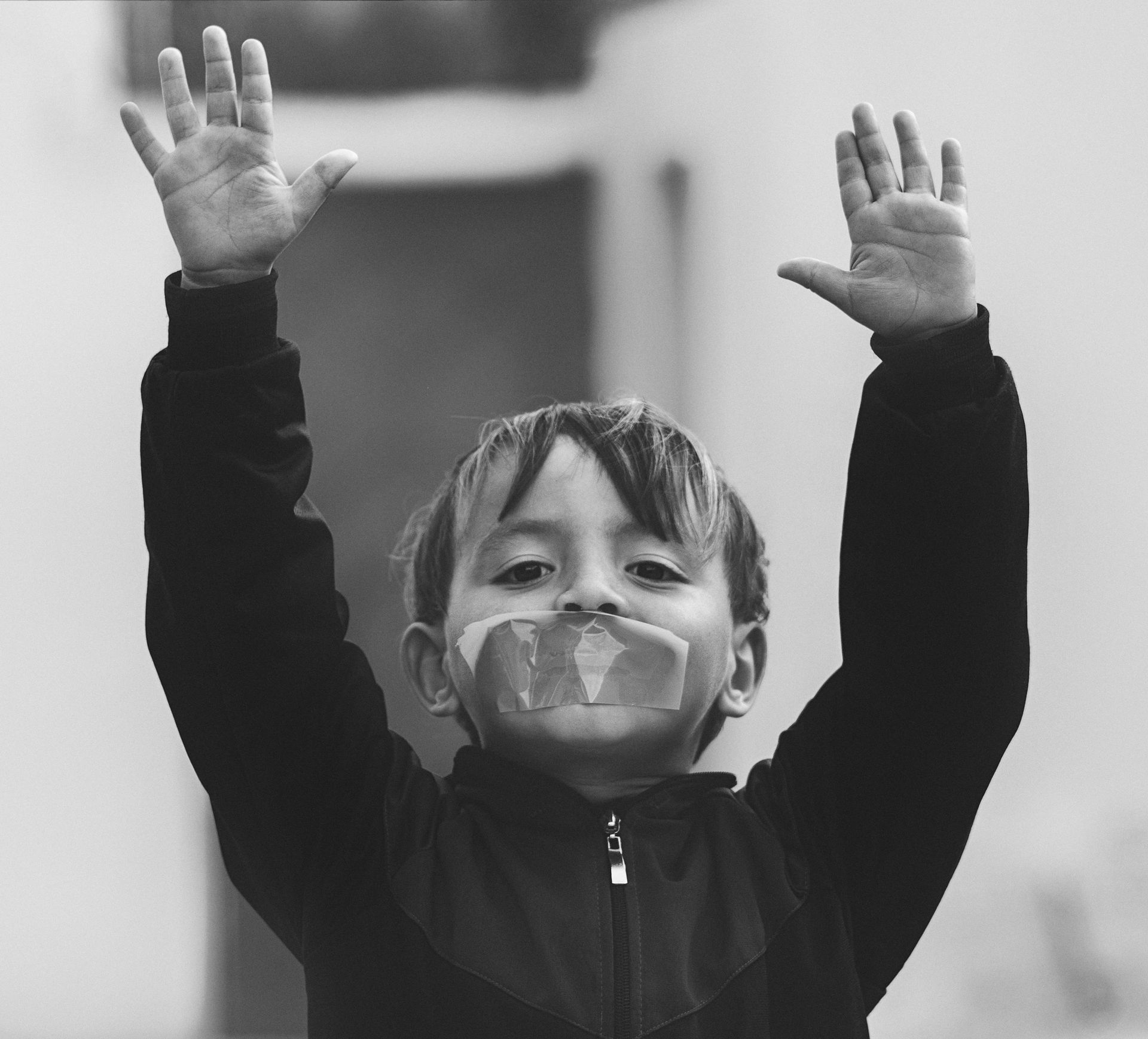 Boy with taped mouth raises hands in the air. Black and white, outdoors.