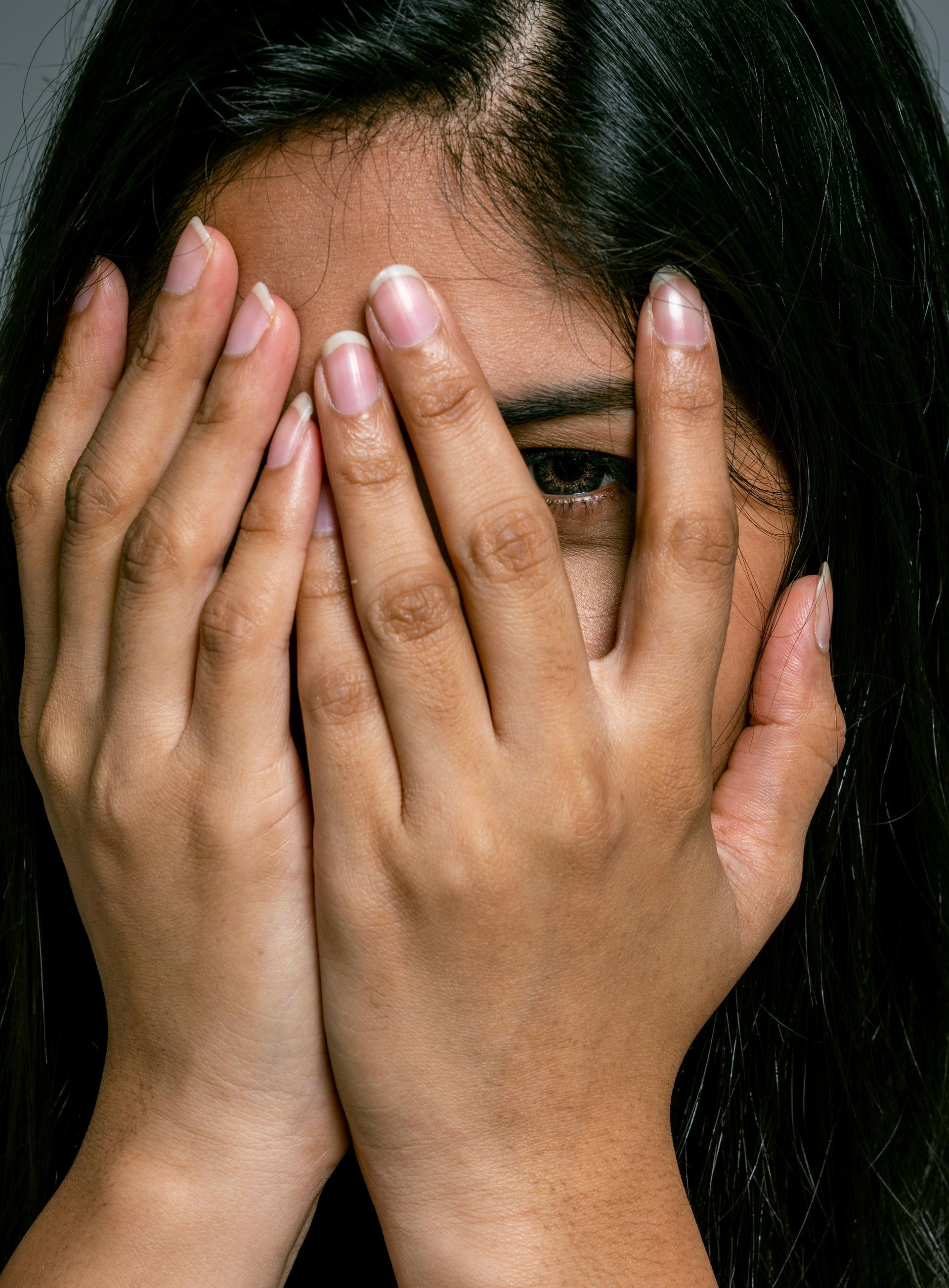 Woman with dark hair covering her face with both hands, eyes peeking through.