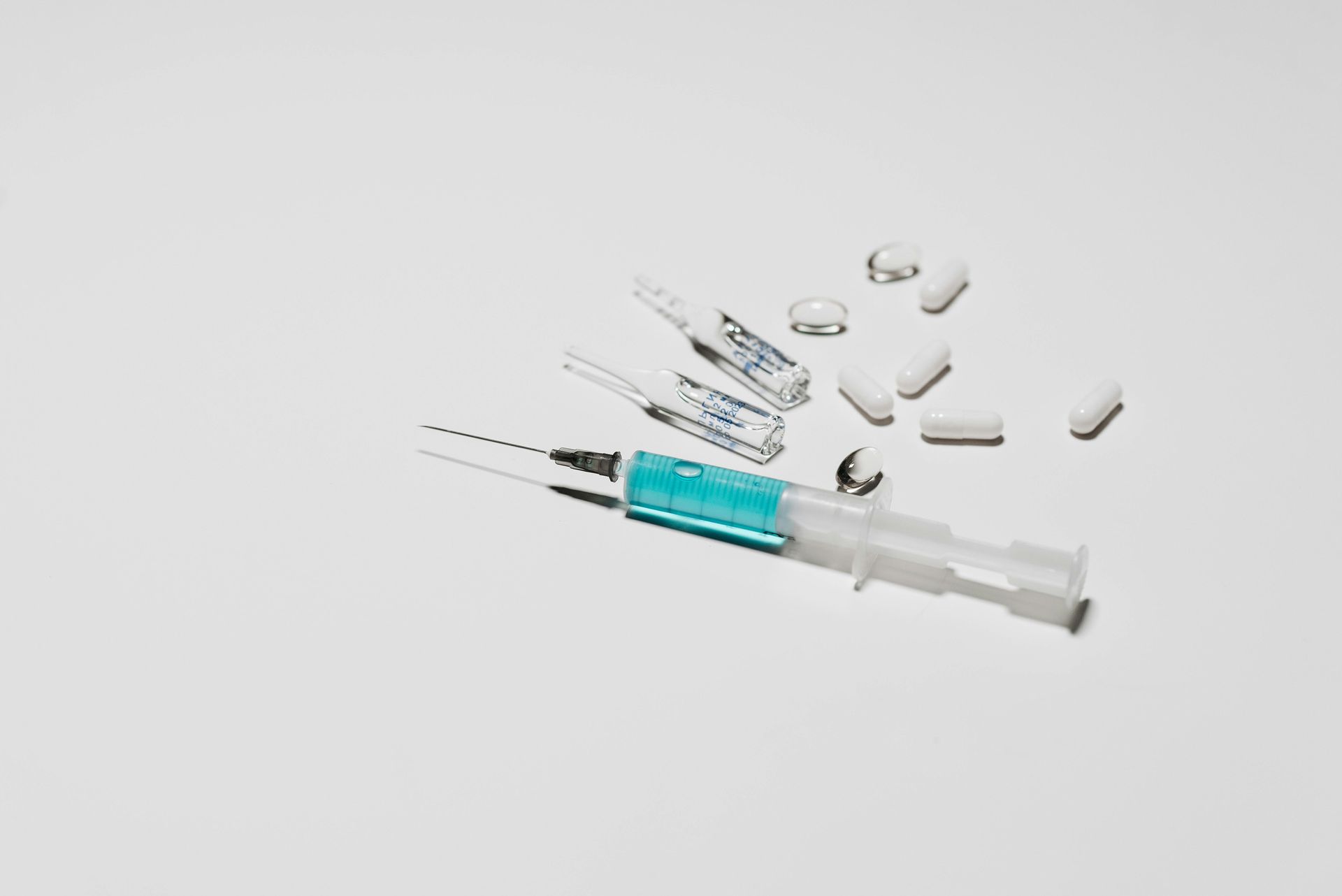 Syringe with blue liquid, vials, and white pills on a white surface.