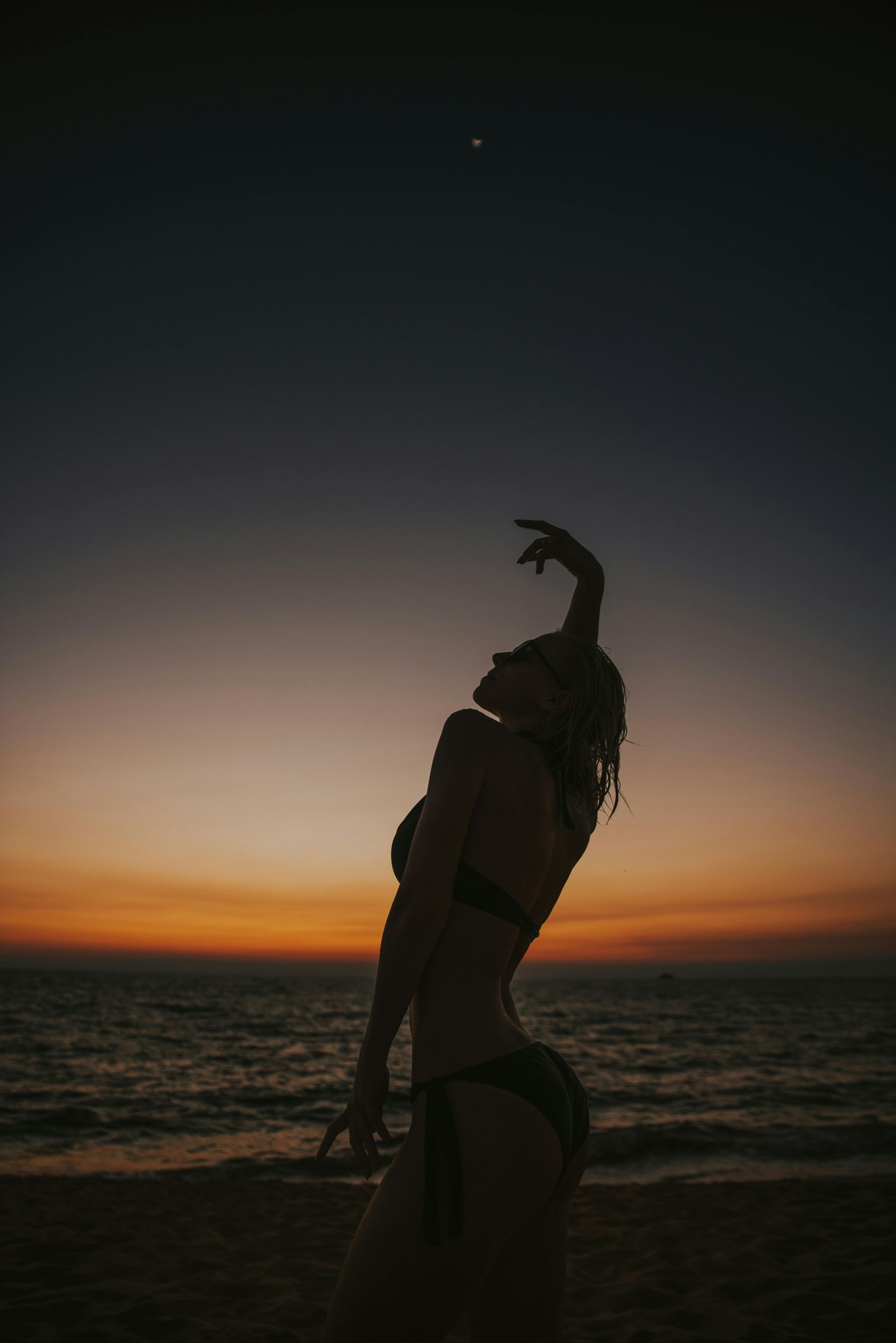 Woman in silhouette on a beach at sunset, reaching up to the sky.