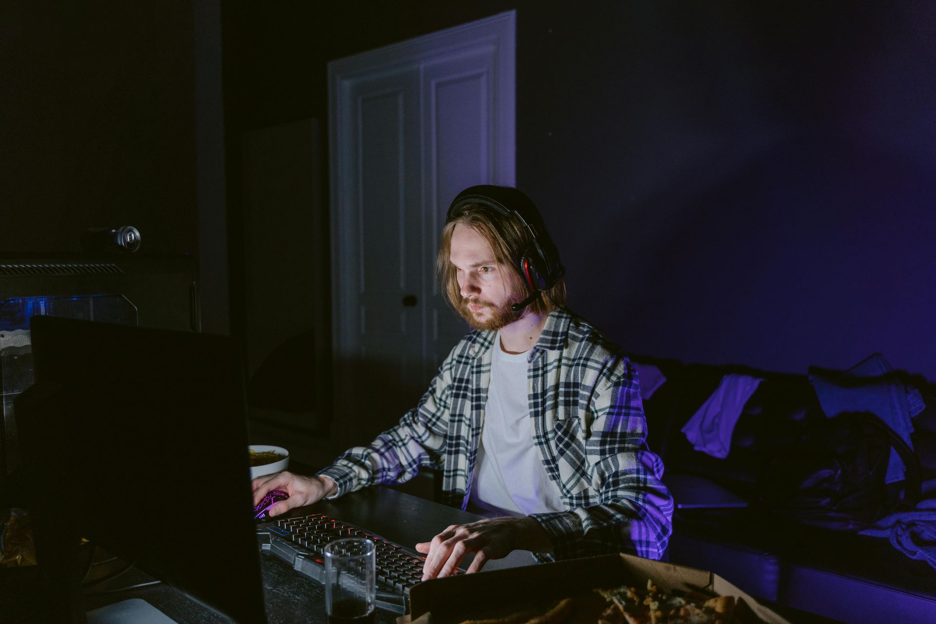 A man with blond hair, wearing a plaid shirt, types on a keyboard in a dimly lit room with a computer.