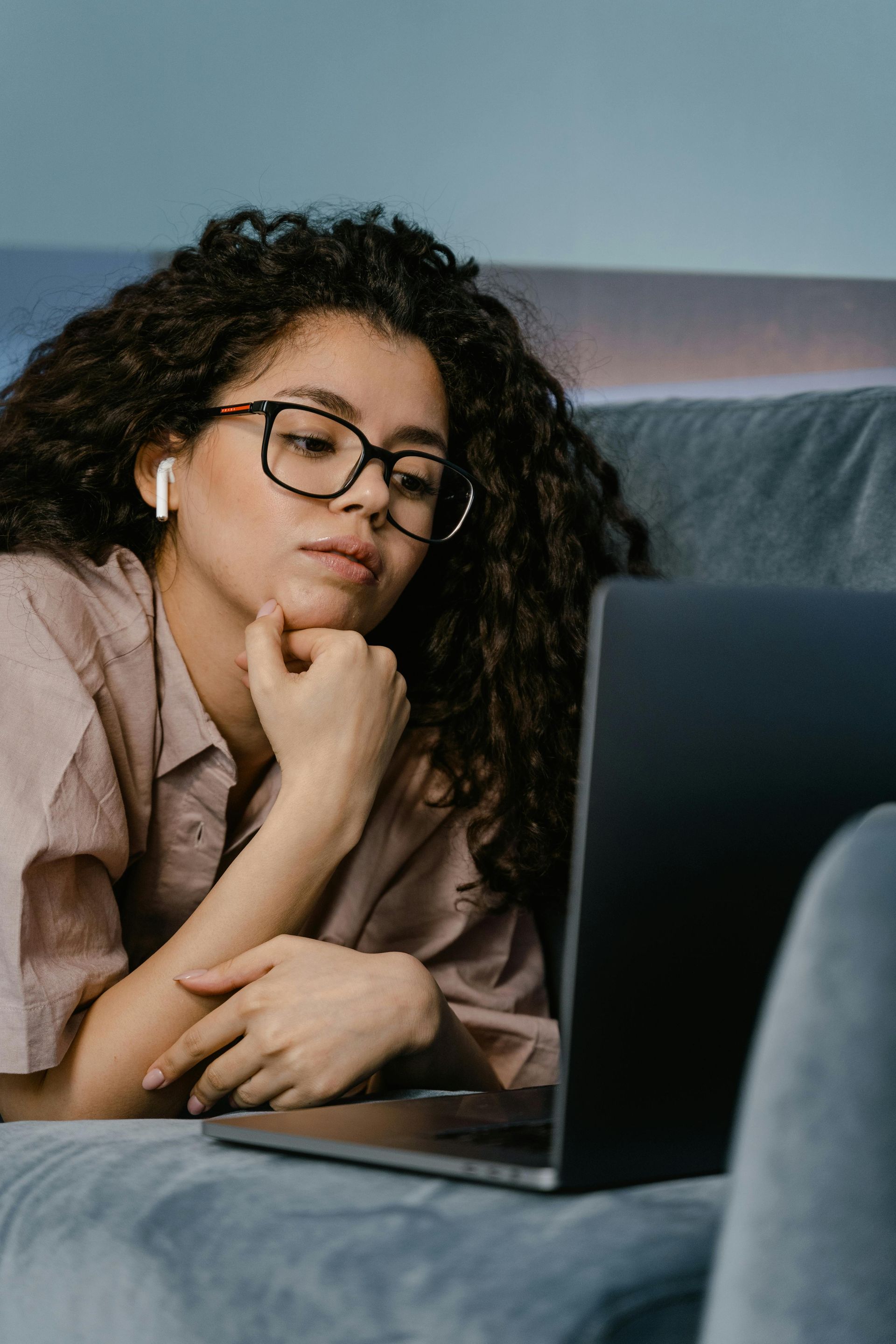 Woman with curly hair, glasses, and earbuds, using a laptop while lying on a couch.