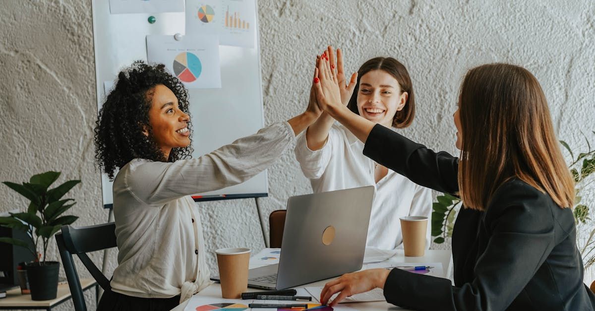 Three women are giving each other a high five while sitting at a table with laptops.
