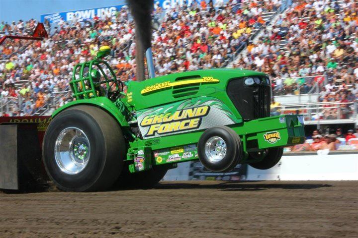 Green John Deere tractor in a tractor pull competition, pulling a weighted sled, with black smoke billowing.