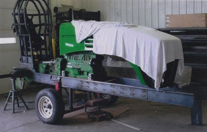 A partially assembled green tractor on a metal frame trailer. Covered in a white sheet, inside a workshop.
