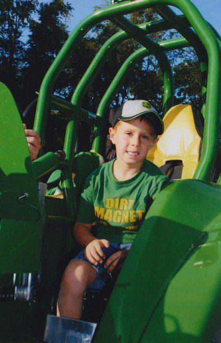 Boy in a John Deere hat and t-shirt sits in a green tractor play structure, smiling.