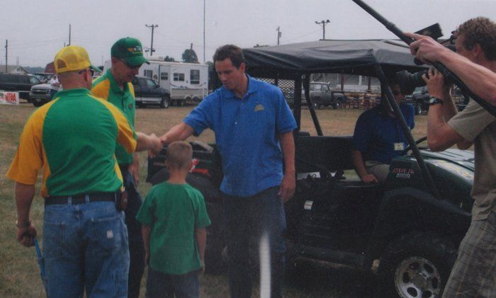 Men in green and blue shirts shake hands with a young boy. A person films with a camera.