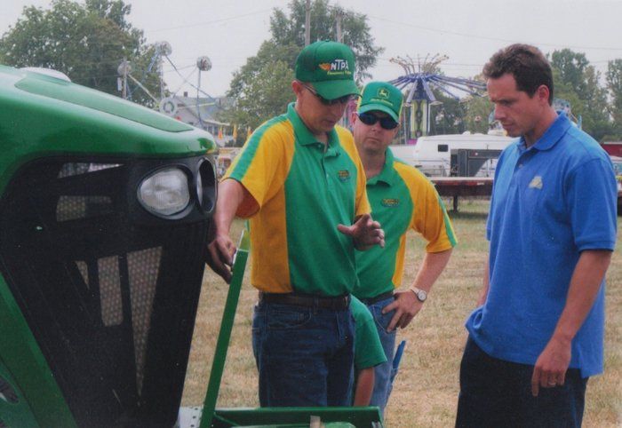 Three men inspecting a green tractor at an outdoor fair. Two men in green/yellow shirts and a man in blue.